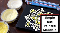 In this image there is a small black canvas on which is painted a dotted mandala, next to the canvas there are three small acrylic containers and on top a number of dotting tools