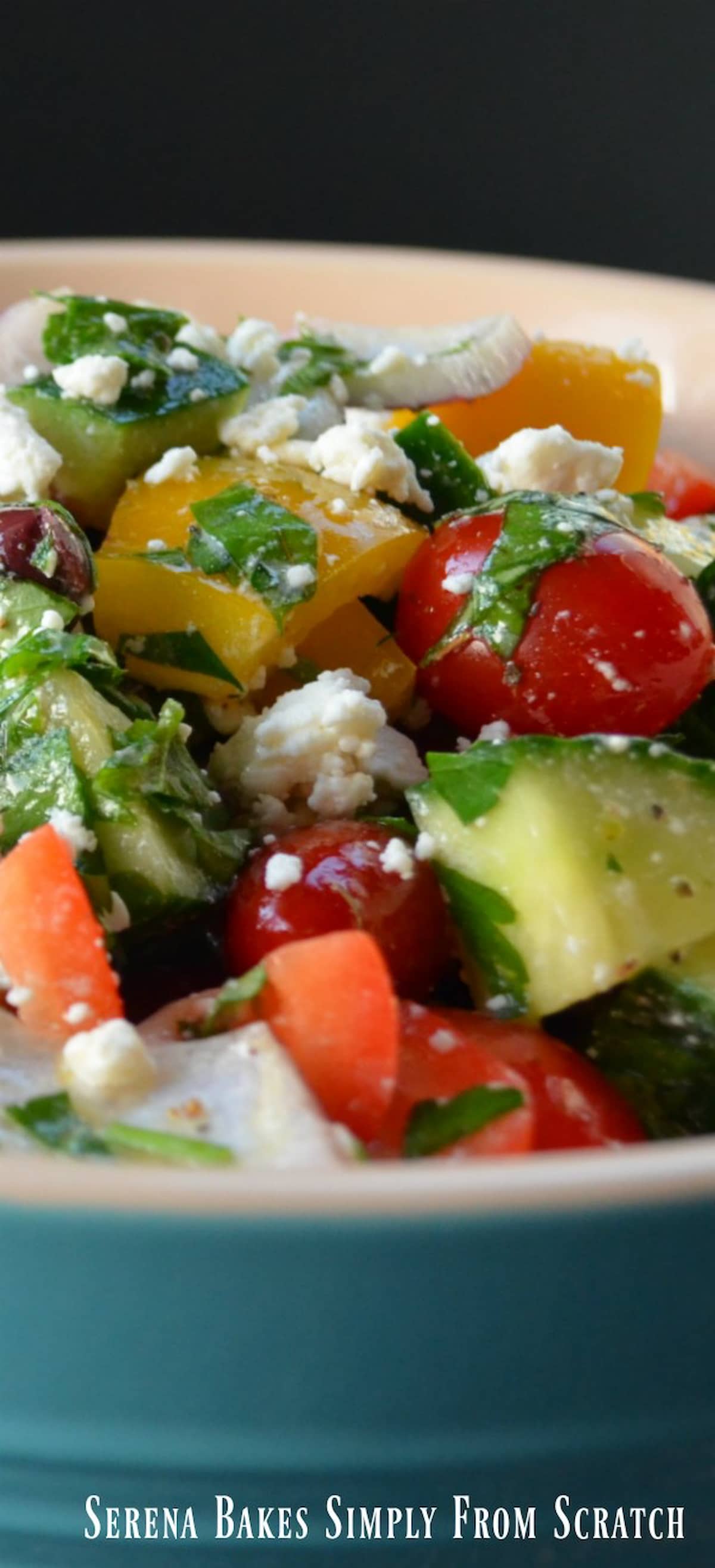 A cream colored bowl on the inside and turquoise on the outside filled with Greek Salad with cherry tomatoes, chopped cucumber, chopped bell pepper, kalamata olives, parsley, and sprinkled with feta cheese.