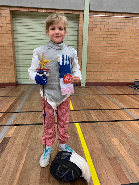 This young one stands smiling proudly holding her new fencing glove and MFC Club patch (a round orange one with purple border and MFC in white). She ’s dressed to fence electric foil, holding her foil in her right hand, tip pointing down. Her fencing mask rests at her feet on the wooden floor of the brick fencing hall.