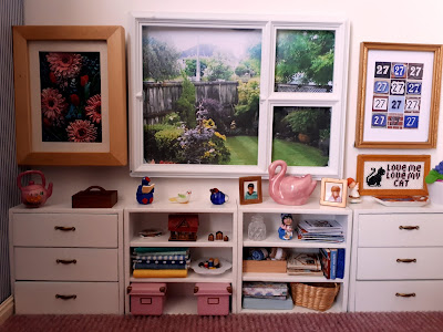 One twelfth scale modern miniature craft room wall with a window overlooking the backyard. Under the window is a row of white bookcases and drawers containing various magazines, books, ornaments and craft supplies. On the wall are various handcrafted pictures.