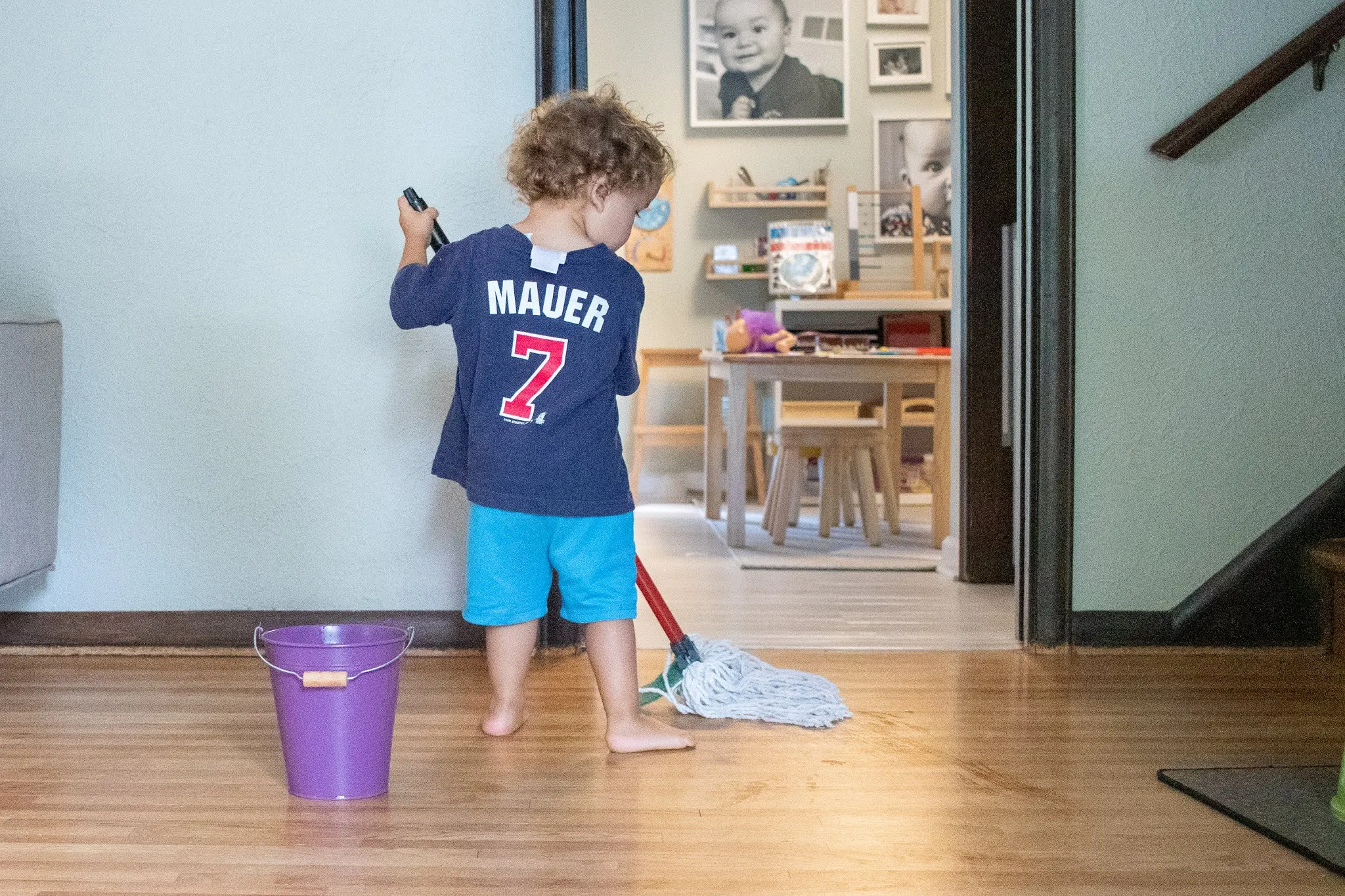 Montessori 2-year-old stands with back to camera near a small bucket of water. In his hands is a child sized mop which he is using to clean the floor.