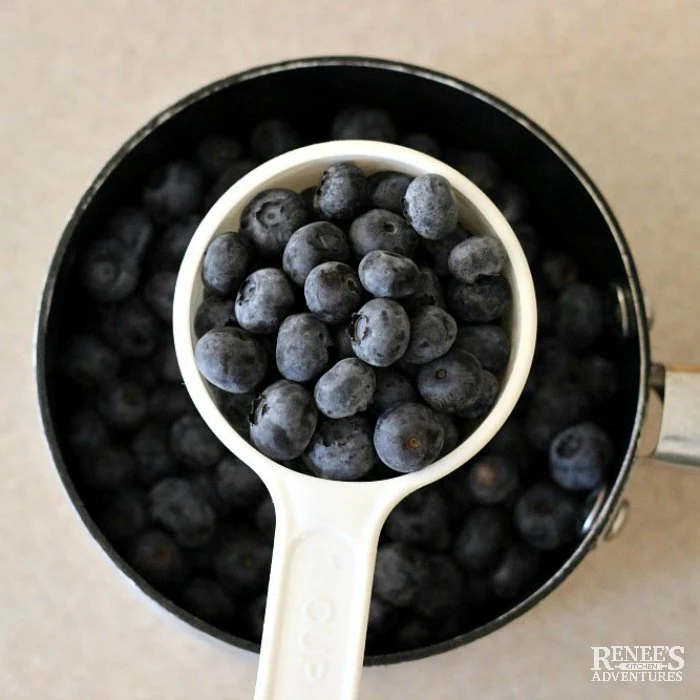 Over head shot of fresh blueberries in a white measuring cup over a pan of fresh blueberries for Blueberry Cream Cheese Pie by Renee's Kitchen Adventures