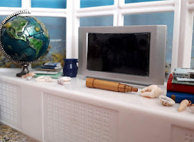 1/12 scale modern miniature windowsill over looking the sea. On the windowsill is a globe, a flat-screen TV, a vintage telescope, several piles of books and a number of shells.