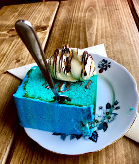 A triangular piece of light blue cake with blue icing with a hemispherical scoop of white ice cream with brown chocolate shards on a blue plate on a rectangular light brown table on a bright background 