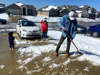Chance, Hannah and TJ, all holding shovels, work to clear the snow and ice covered driveway. A white Prius sits at the end of the driveway.  Surrounding homes have melting snow on the roofs.