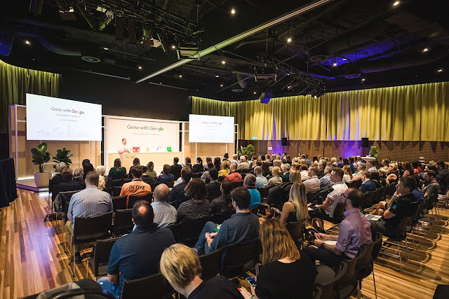 People sit in rows of chairs in a conference room, with a presentation on stage about Grow with Google