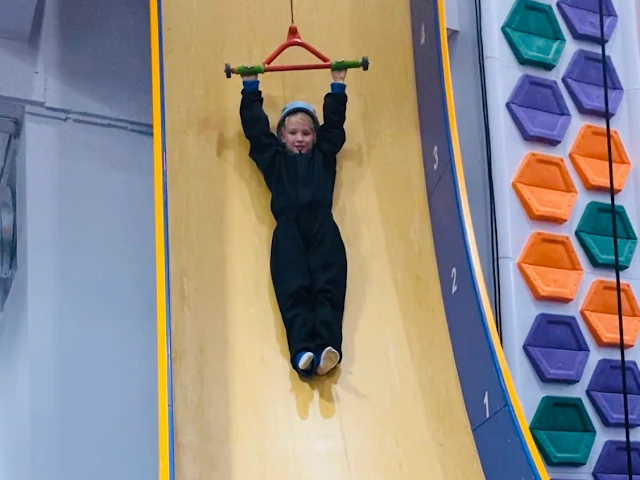 A girl in a helmet and black boiler suit holding on to a bar which is pulling her vertically up a slide