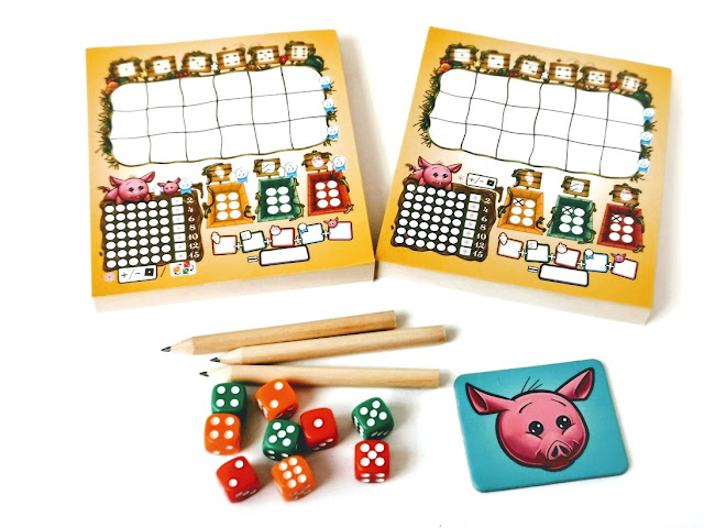 Kości obfitości roll&write dla dzieci,na zdjęciu elementy gry: dwa bloczki do zapisywania wyników, pod nimi leża małe ołówki, dziewięć kolorowych kostek i znacznik gracza rozpoczynającego z wizerunkiem głowy świnki na niebieskim tle