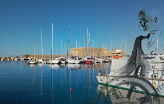 Παρουσιάζεται Το παλαιό, βενετικό, λιμάνι του Ηρακλείου, στο φόντο το επιθαλάσσιο οχυρό Rocca a Mare / Κούλες, ενώ στο λιμάνι διακρίνονται καΐκια και ιστιοπλοϊκά 