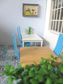 Modern dolls house miniature cafe scene, with blue and white tiled floor, white and grey wallpaper, white and beech tables, blue chairs and plants in the foreground.