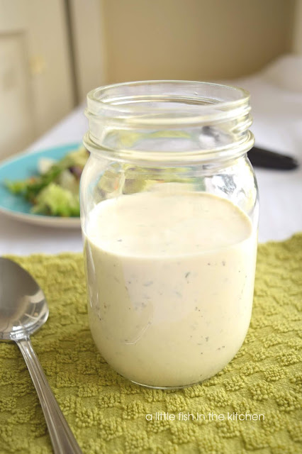 A clear mason jar filled with creamy, white ranch dressing is sitting on a green textured kitchen towl. A spoon rests beside the jar and green salad on a blue plate is slightly blurred in the background behind the jar of salad dressing. 