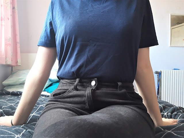 The image shows a model showing the Goose Studios Navy T-shirt - a sustainable sourced piece of clothing. It is paired with a black ripped jeans. The model is sitting on a black blanket, on a bed.