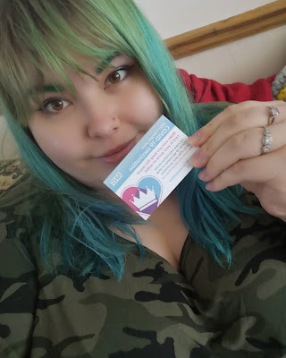 Selfie of Nici with green hair, camouflage top, holding up vaccination card.