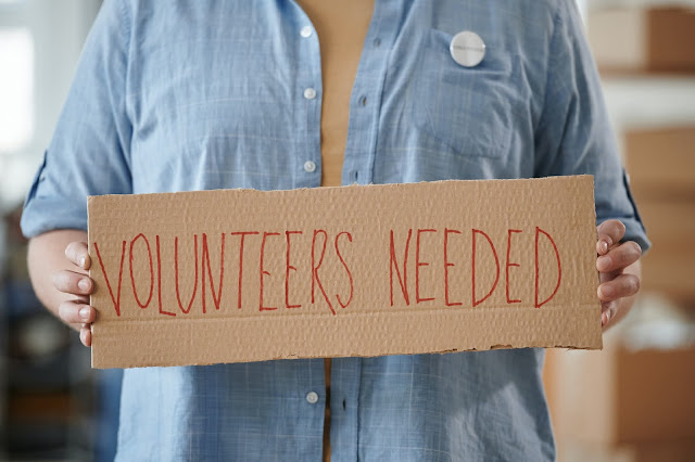 A cardboard sign with the text "VOLUNTEERS NEEDED" written on it.