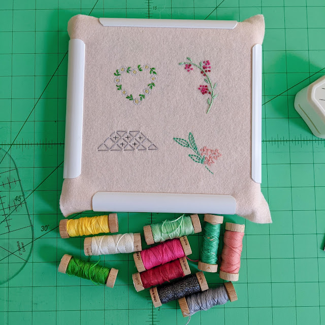 A square embroidery frame holding felt with 4 embroideries, 3 floral and one geometric, with lots of spools of Aurifloss below it