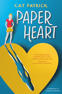 A girl wearing shorts with her hands in her pockets standing on top of a large paper heart that is torn into two large pieces