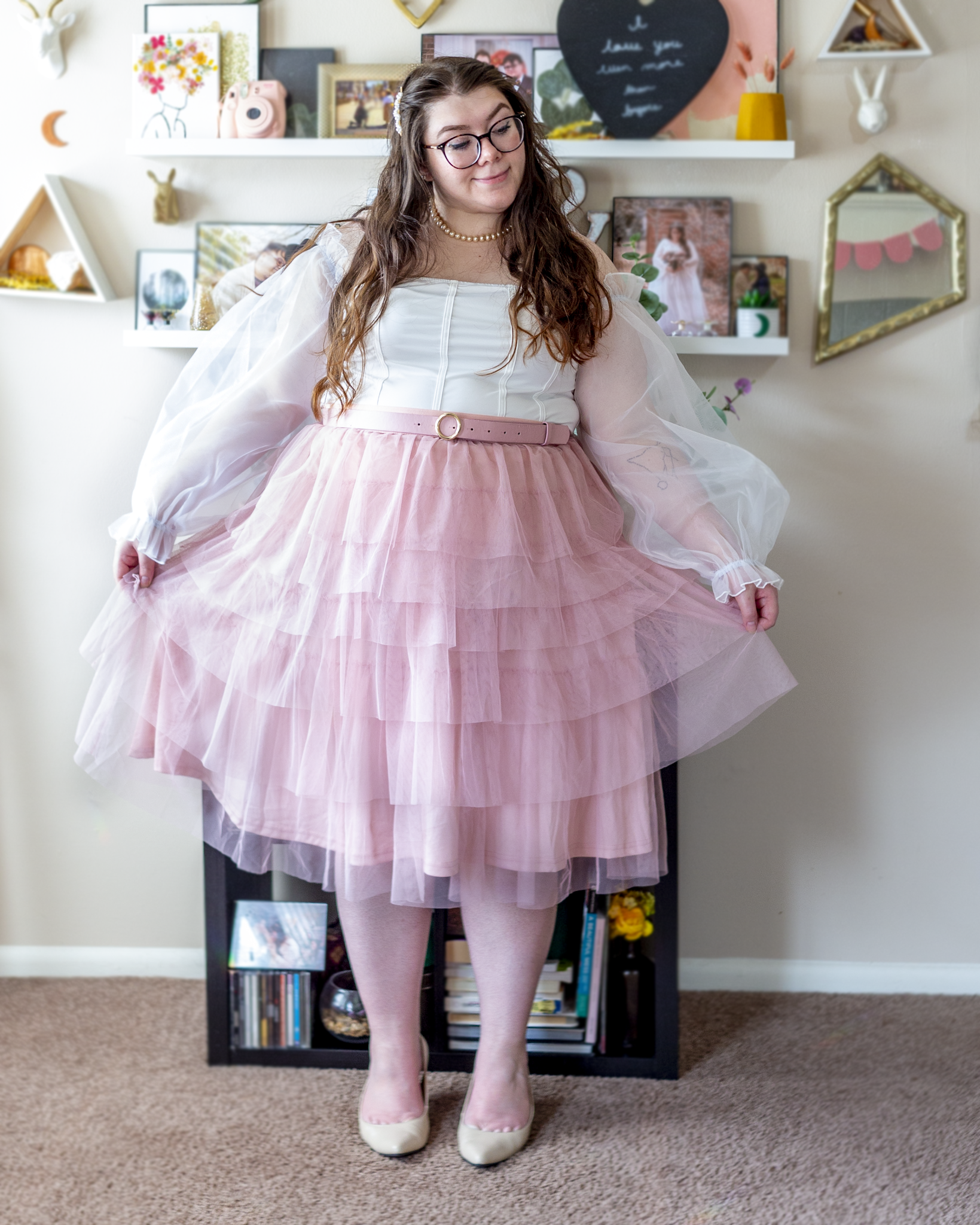 An outfit consisting of a white corset inspired bodysuit with off the shoulder long sheer lantern puffy sleeves with a pastel pink tiered tulle midi skirt layered over, belted with a coordinated pink belt and beige pointed toe slingback heels.