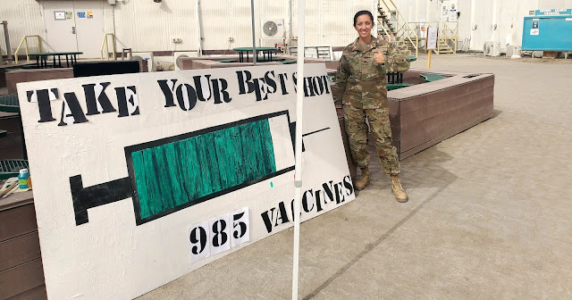 Jeanmarie Rey in front of a big sign that has a needle and says "TAKE YOUR BEST SHOT - 985 VACCINES"