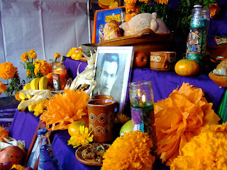 An ofrenda A view of a colorful ofrenda. There is a black and white portrait in the middle with many yellow flowers and food on a purple cloth.