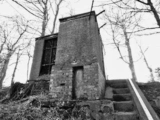 Black and white photograph of the SEPA Monitoring Site, as seen from the riverside.  It is a small building with a large, boarded up window facing out to the river.  There are steps leading down to the river at the side of the building.  Photo by Kevin Nosferatu for the Skulferatu Project