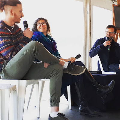 Three people seated on a stage. One is talking into a microphone, while the other two look on, one with micrphone poised to ask a question.