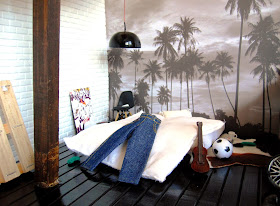 One-twelfth scale modern miniature loft space with a palm tree mural on the wall behind a futon bed with jeans draped over it.