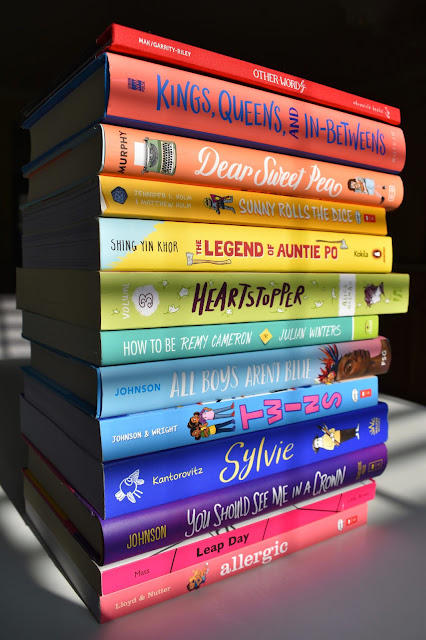 A stack of 13 books with spines in a rainbow pattern from red at the top to pink at the bottom. The spines take up the right three-fourths of the image and are brightly illuminated except for some dark vertical and horizontal stripes from the shades. The tops of the books are visible in the left fourth of the image and are dark. The surroundings are almost completely black, except for a few stripes of light in three locations on the white table.