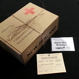 Cardboard box made to resemble a Red Cross box, next to a ration book and a name tag for NZAME convention 2016.