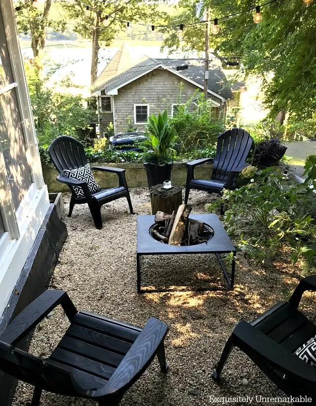 Black Adirondack chairs with white and black pillows  around a fire pit on a gravel patio