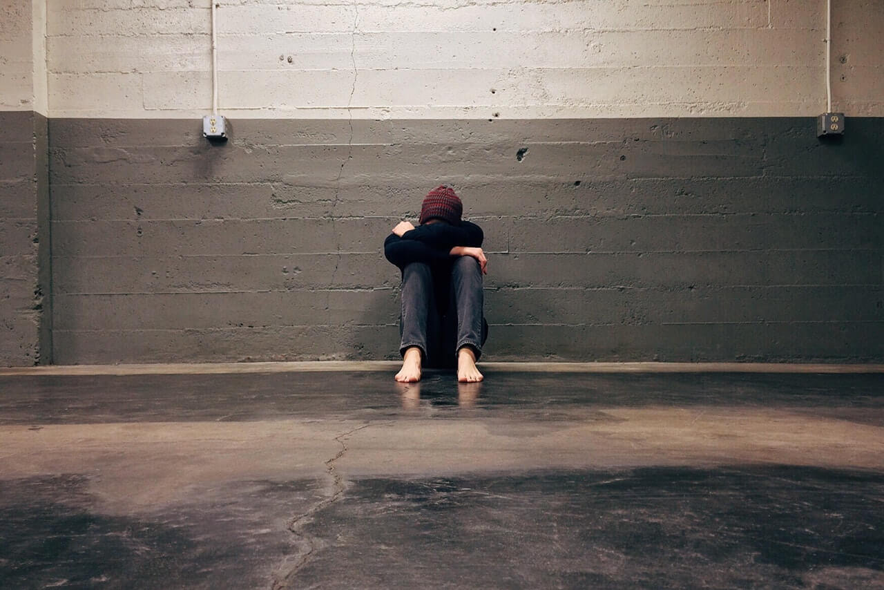 A depressed boy sitting against the wall with head down between the legs.