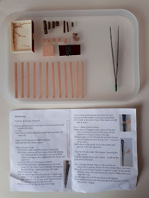 Pieces of a 1/12 scale miniature kit laid out neatly on a small tray next to a pair of tweezers. Next to the tray is a set of instructions for the kit.