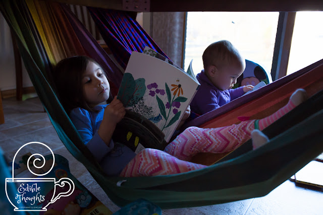 Image of two children reading in wrap hammocks. White watermark in bottom left corner of a steaming mug with a needle horizontal along the rim and curly text Edible Thoughts copyright symbol Stephanie Lu Sinclair.]