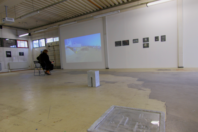a general view of the show taken with a fish eye lens.A video work shows a ferris wheel, with small photographs on the right side, a person sitting in front of the video, and more works far on the left. The floor shows wear and is maybe laminate, tinting towards yellow. a general view of the show taken with a fish eye lens.A video work shows a ferris wheel, with small photographs on the right side, a person sitting in front of the video, and more works far on the left. The floor shows wear and is maybe laminate, tinting towards yellow.