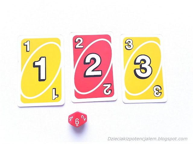 na zdjęciu trzy karty uno o wartościach jeden, dwa i trzy oraz dziesięciościenna kostka w kolorze czerwonym