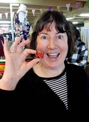 Woman holding up a brooch in the shape of the word 'yay' next to her mouth.