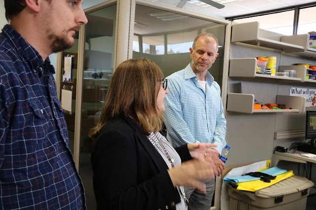 A woman is talking to a man and using her hands to explain. He is studiously looking at her. Another man to the side stares off into the distance. They are in a library.