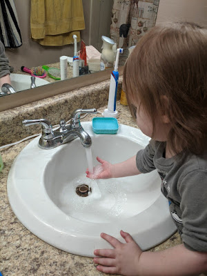 ID: a two-year-old with pale skin, light brown hair, and rolled up sleeves holds a hand under a faucet of running water.