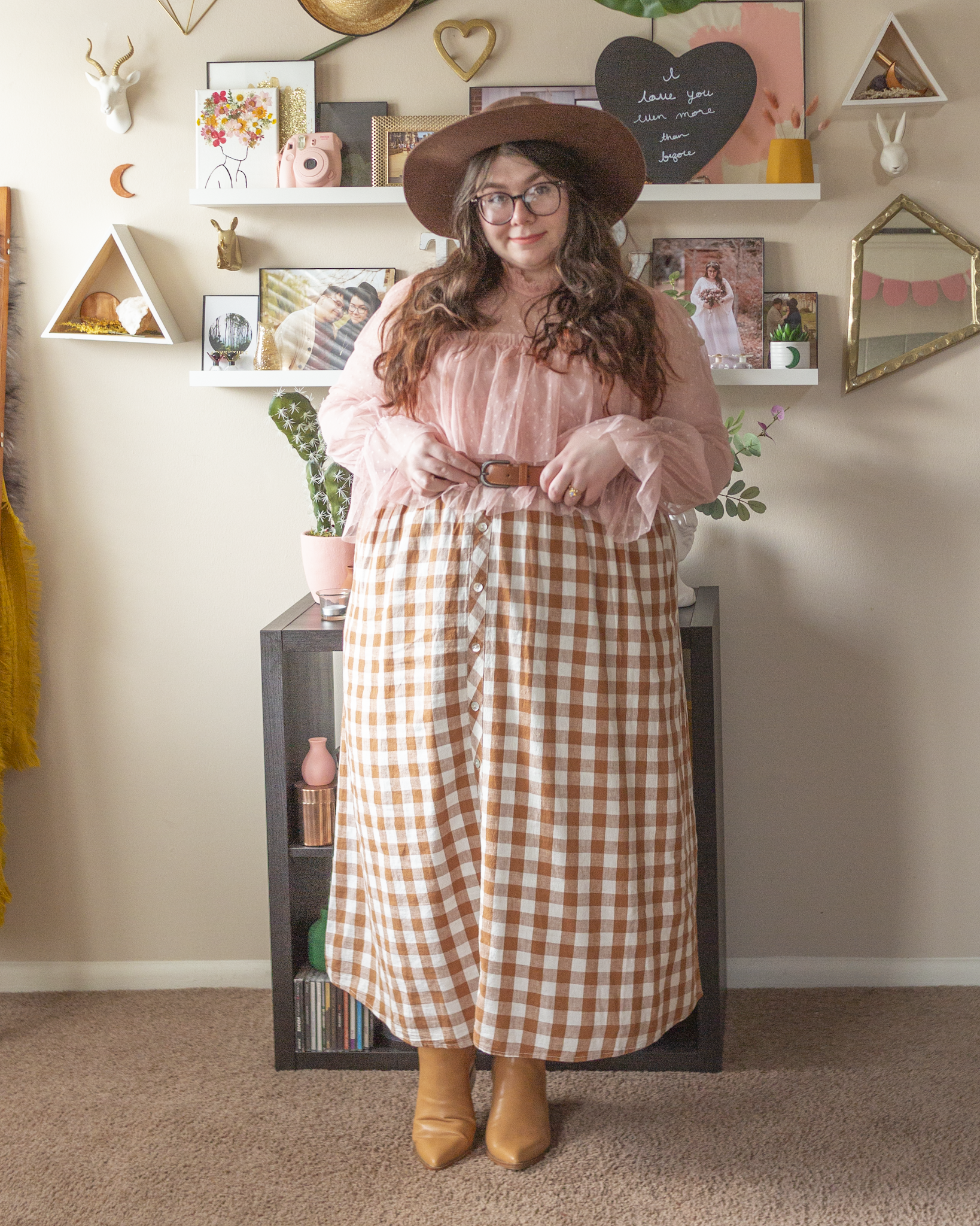 An outfit consisting of a dark brown wide brim hat, pastel pink high neck long sleeve sheer tiered blouse with polka dots and ruffle hem sleeve tucked into a brown and white gingham button down dress, worn as a skirt, and camel brown pointed toe Chelsea boots.
