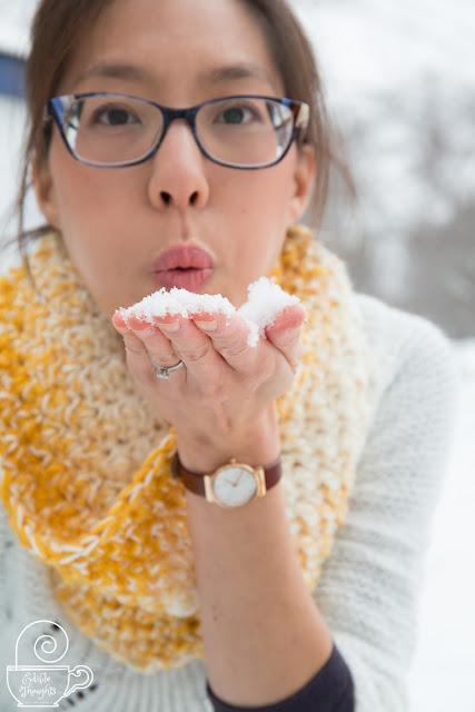 Image of a tan skin bespectacled Asian woman blowing snow from her bare palm towards the camera. She's wearing a yellow and cream crocheted cowl over a white sweater. Behind her is a winter wonderland. 