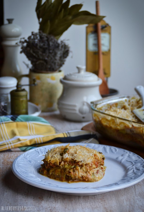 Lasaña de berenjenas en fuente de cristal sobre tabla de madera, con ingredientes de fondo, hierbas aromáticas, laurel seco, molinillo pimienta