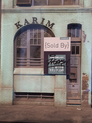 Detail of the front of a 1/24 scale model facade of an old building with the word "KARIM' above the window and a grafittied 'sold' sign on the wall.