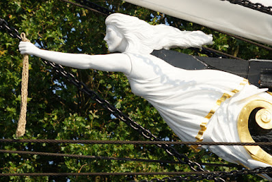 Photograph of a carved white figurehead of a woman in a shift dress holding a horse's tail in her outstretched hand.