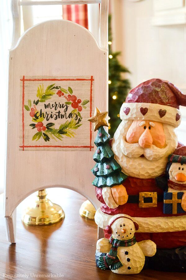 Small wooden sled decorated with Merry Christmas napkin next to Santa on table