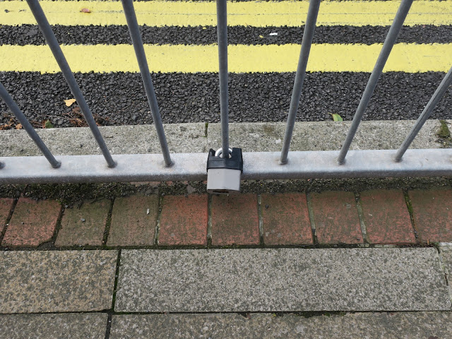 Lone padlock fixed to railings by yellow lines and brick edged pavement.