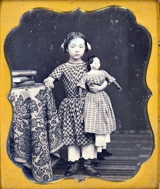 Fotografía post mortem sobre 1850.  Niña y muñeca, vestido a cuadros y pantalones por debajo.  https://highlandscurrent.org/2015/04/07/daguerreotype-exhibit-at-vassar-opens-april-10/