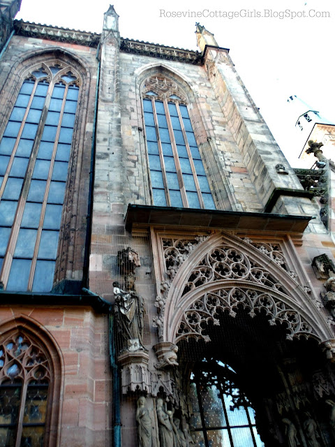 Photo of the amazing carvings above one of the doors of a church in Nuremberg Germany called St. Sebald church.  Above the doorway are towering windows  The lower window to the left of the door is also large. All of the windows have carved wood swirls and other features. Photo by rosevinecottagegirls.com
