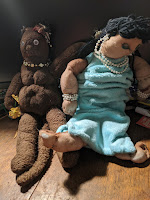 A brown velvet cat wearing a beaded necklace seated next to a doll with black hair and a light blue velvet dress. Do not forget the kind of hooked feet. Good thing this doll does not have to stand up.