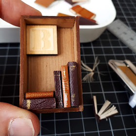Hand holding a miniature wooden box frame containing a wooden 'B' tile, and five light and dark brown books stacked inside. In the background is a cutting mat, retractable knife, rulerm container of miniature books and some pieces of cut books.