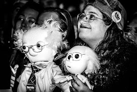 Black and white photo of a white woman wearing glasses, looking upward and smiling, holding two Muppet-like dolls