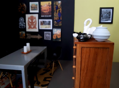 One twelfth scale modern miniature dining room with black walls hung gallery style, an Eames dining chair and yellow rug with the number 7 on it in black. In the foreground is a tall chest of drawers with a white china swan and vase on it